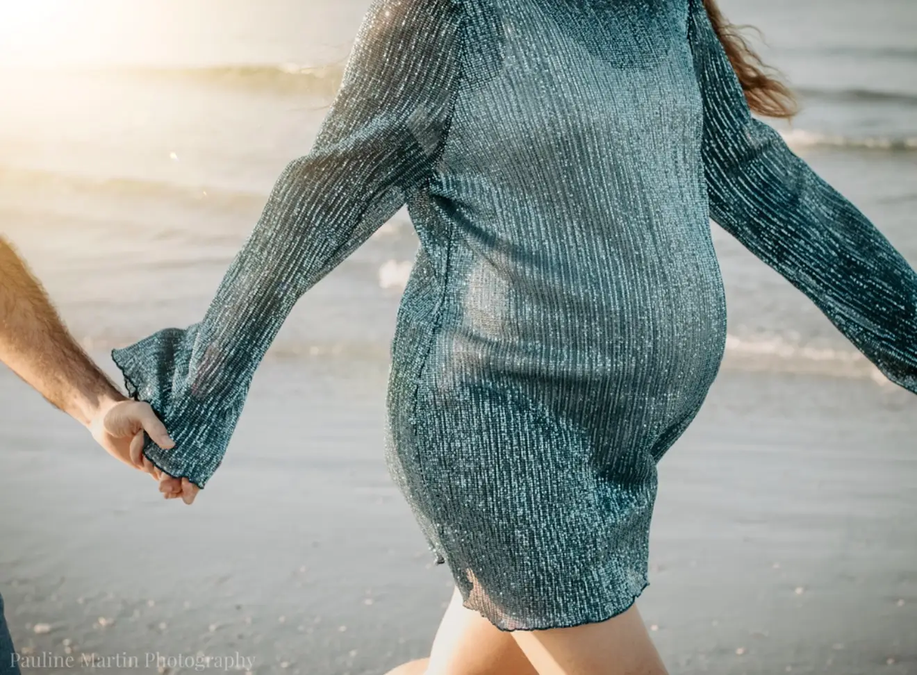 Séance grossesse en bord de mer : gros plan sur le ventre et sur les mains du couple qui se tiennent, à contre-jour.
