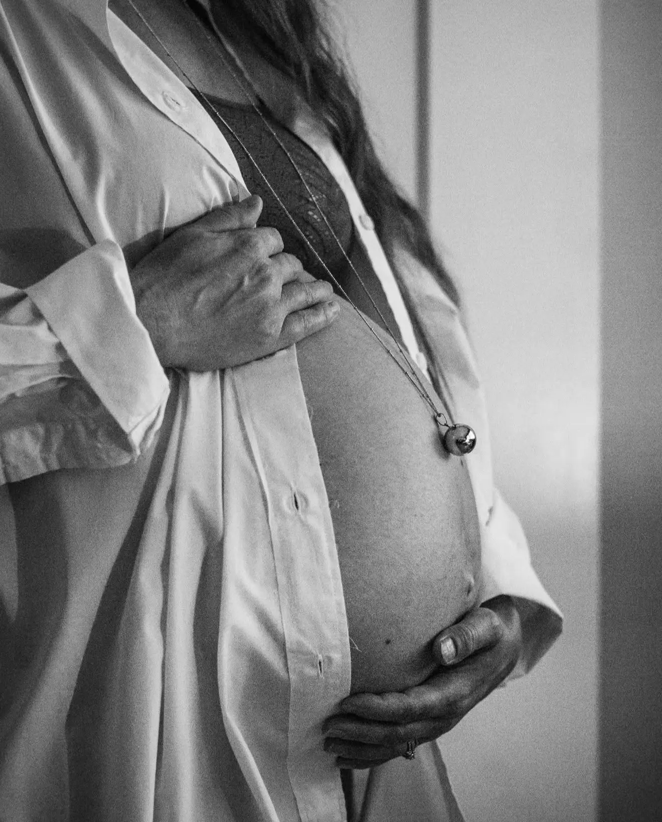 Séance grossesse à domicile : les mains du futur papa posées sur le ventre rond, bola de grossesse autour du cou, en noir et blanc.