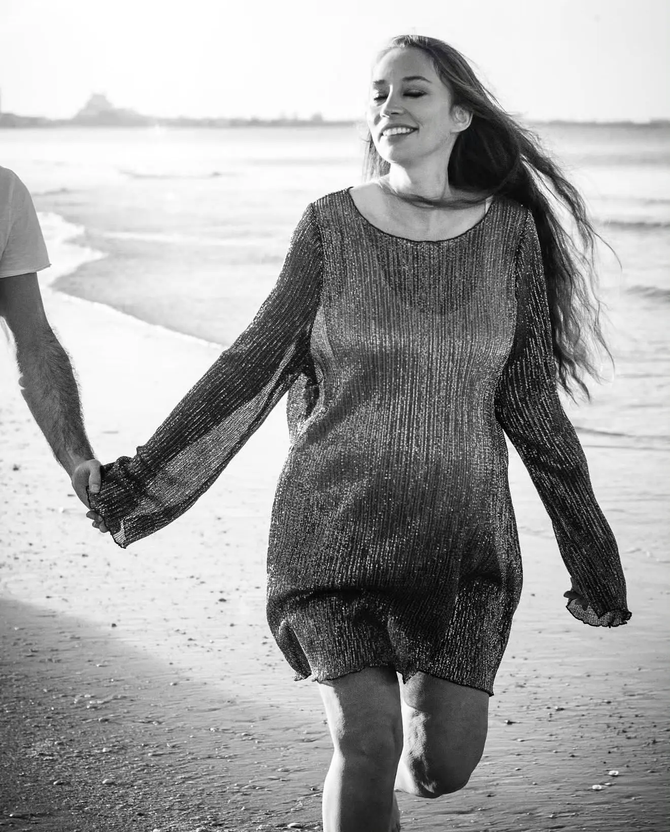 Séance grossesse sur la plage : future maman courant sur le sable, main dans la main avec son compagnon, en noir et blanc.