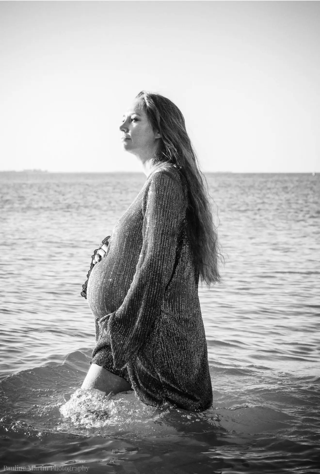 Séance grossesse les pieds dans l'eau : future maman de profil, la mer jusqu'aux genoux, en noir et blanc.