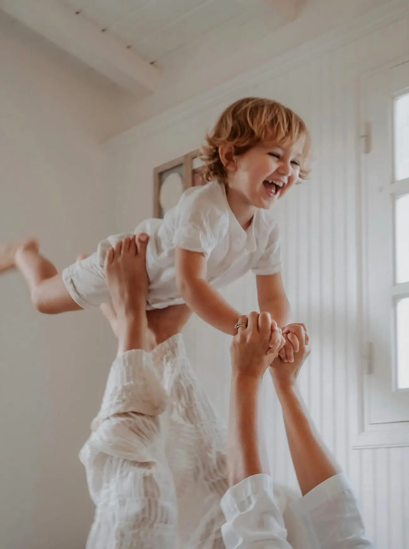 Séance famille à domicile, au Gosier : un enfant soulevé en avion au-dessus de sa maman, en plein éclat de rire, dans une pièce toute blanche.
