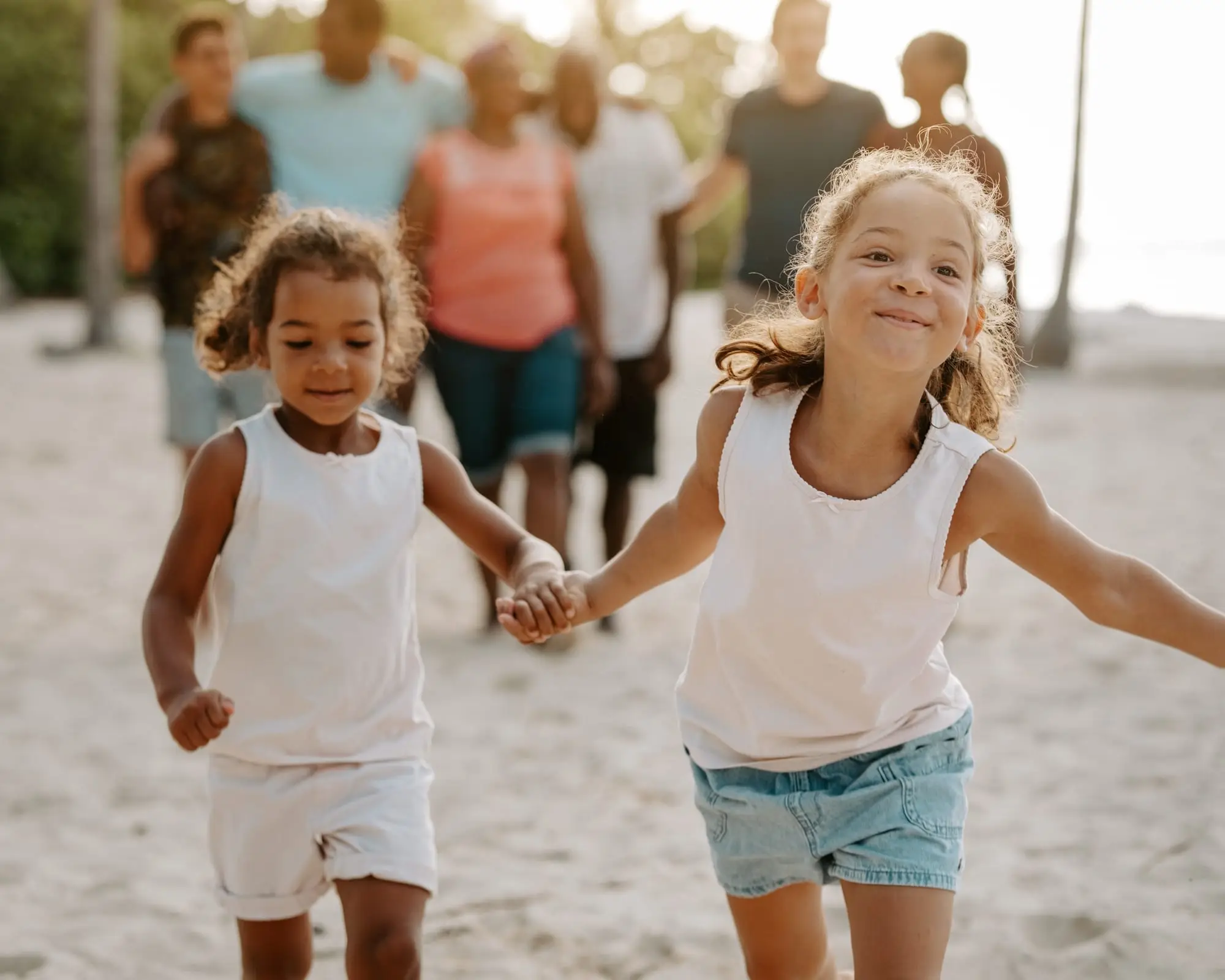 Séance famille en Guadeloupe : deux petites filles courent main dans la main sur la plage de Bois Jolan, à Sainte-Anne, le reste de la famille avançant derrière elles dans la lumière dorée du matin.