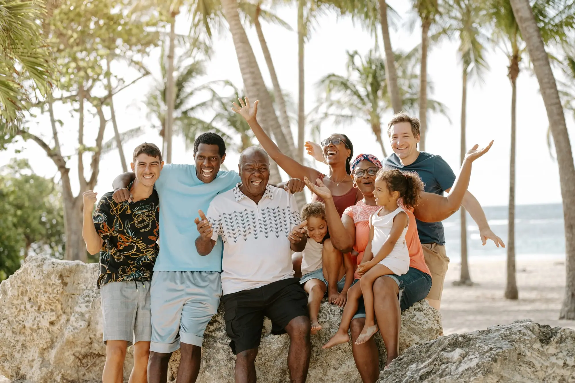 Séance famille sur la plage de Bois Jolan, à Sainte-Anne : les huit membres de la famille réunis sur un rocher sous les cocotiers, bras levés, la mer derrière eux.