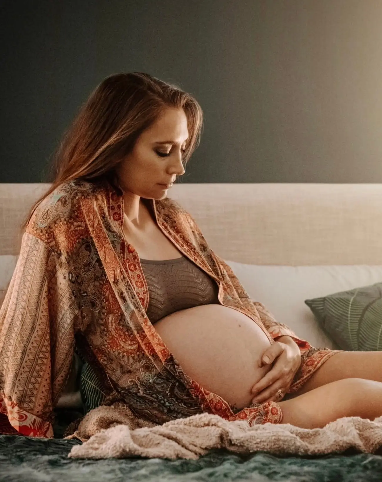 Séance grossesse à domicile : future maman assise sur son lit en kimono paisley, les mains posées sur le ventre.