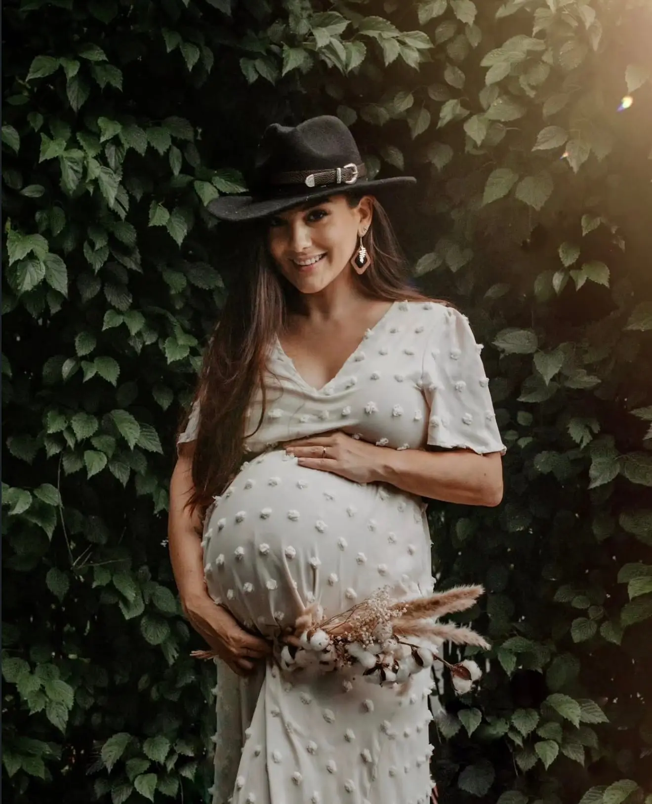 Séance grossesse en extérieur : future maman au chapeau noir devant un mur de feuillage, un bouquet de fleurs séchées contre le ventre.