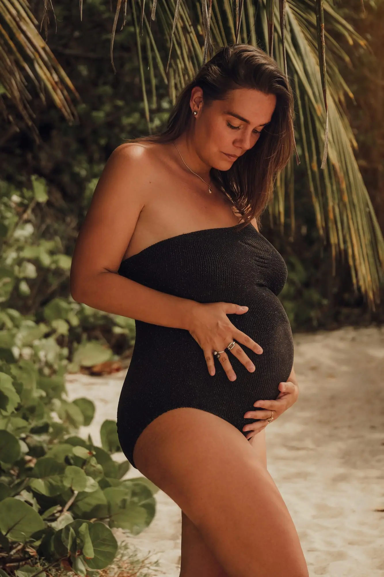 Séance grossesse sur la plage de l'Anse Colas, à Anse-Bertrand : future maman en maillot noir sous un palmier, les deux mains sur le ventre rond.