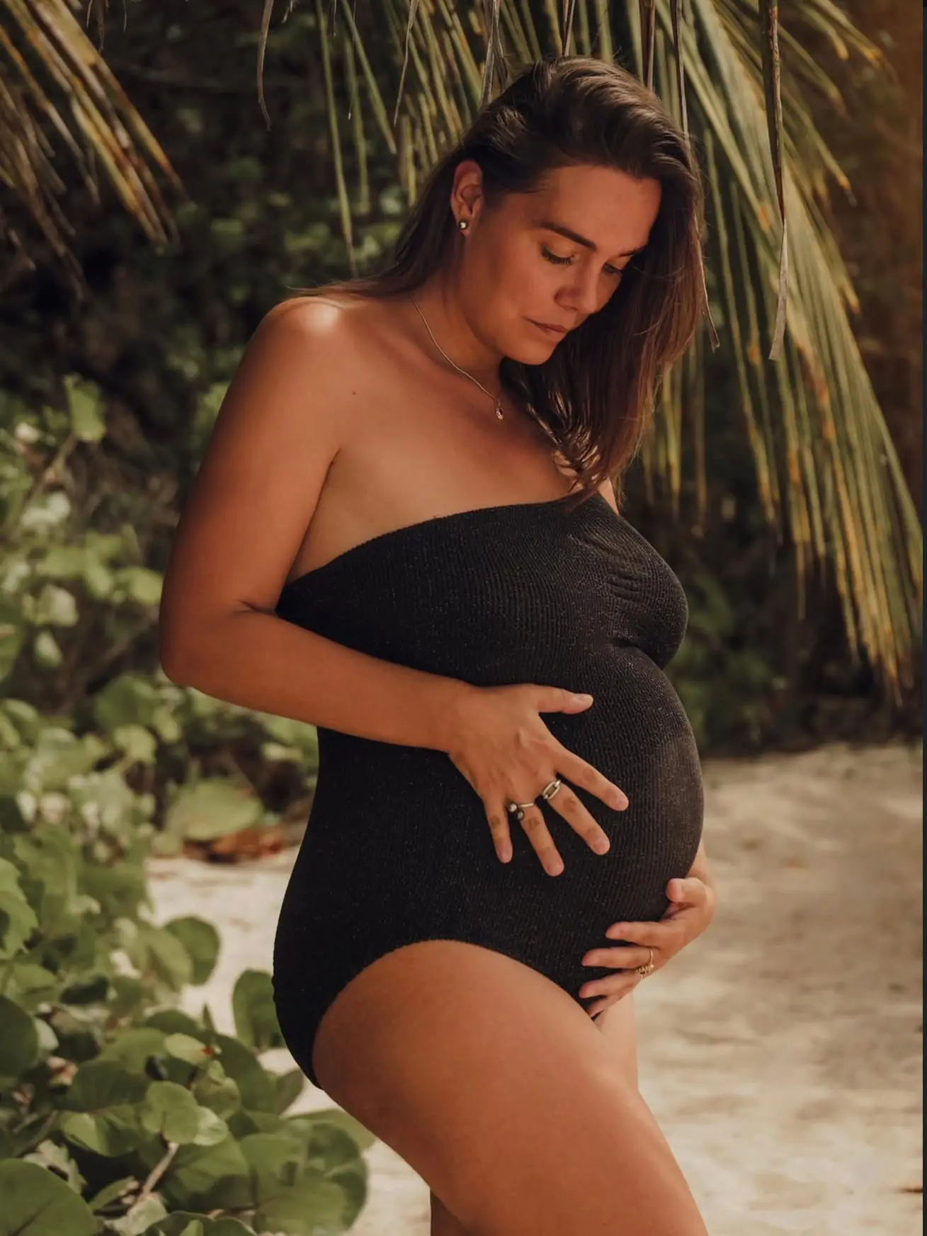 Séance grossesse sur la plage de l'Anse Colas : future maman en maillot noir sous un palmier, les deux mains sur le ventre rond.