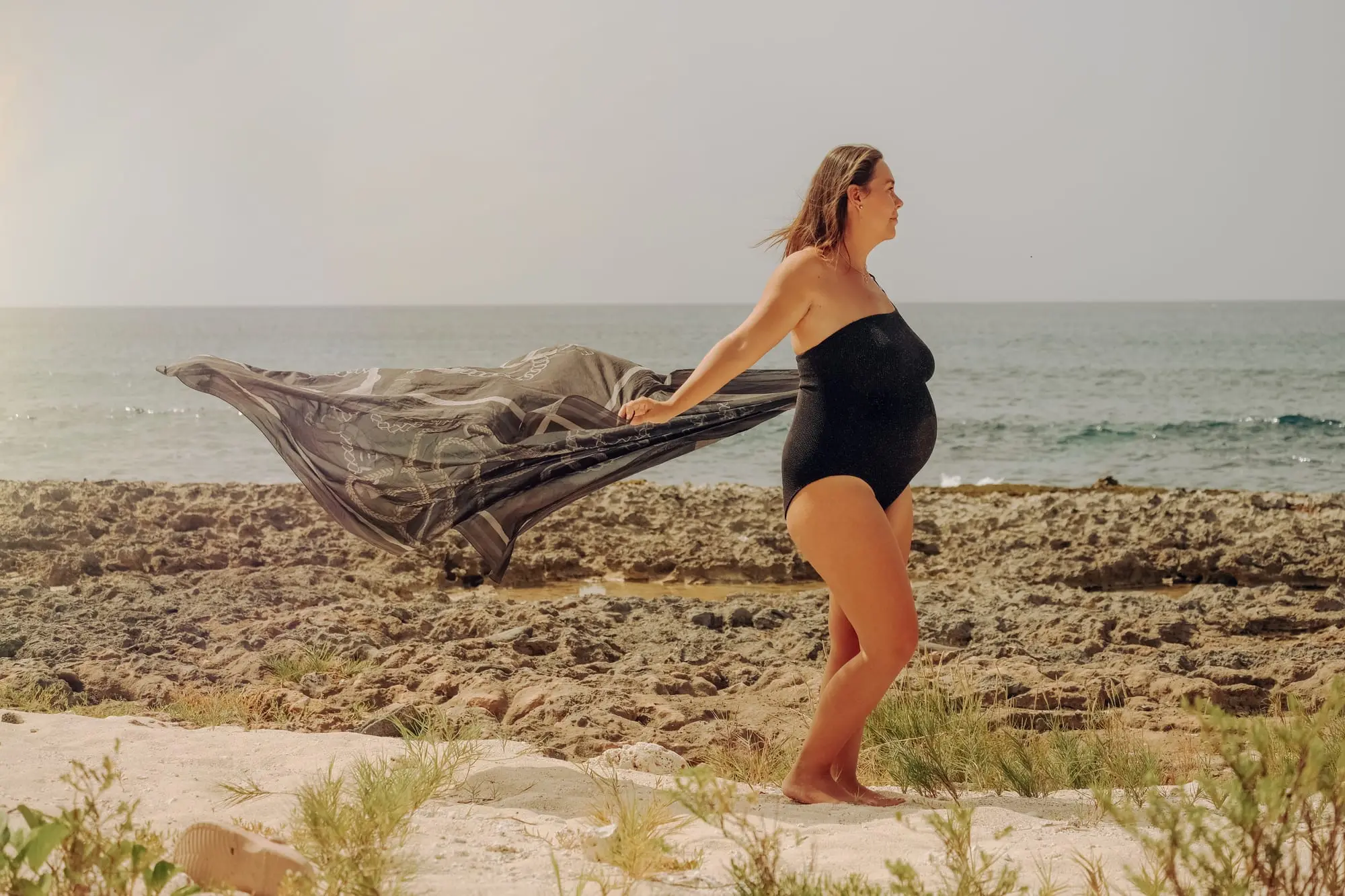 Séance grossesse sur la côte sauvage de l'Anse Colas, à Anse-Bertrand : future maman en maillot noir, son paréo soulevé par la brise, la mer derrière elle.