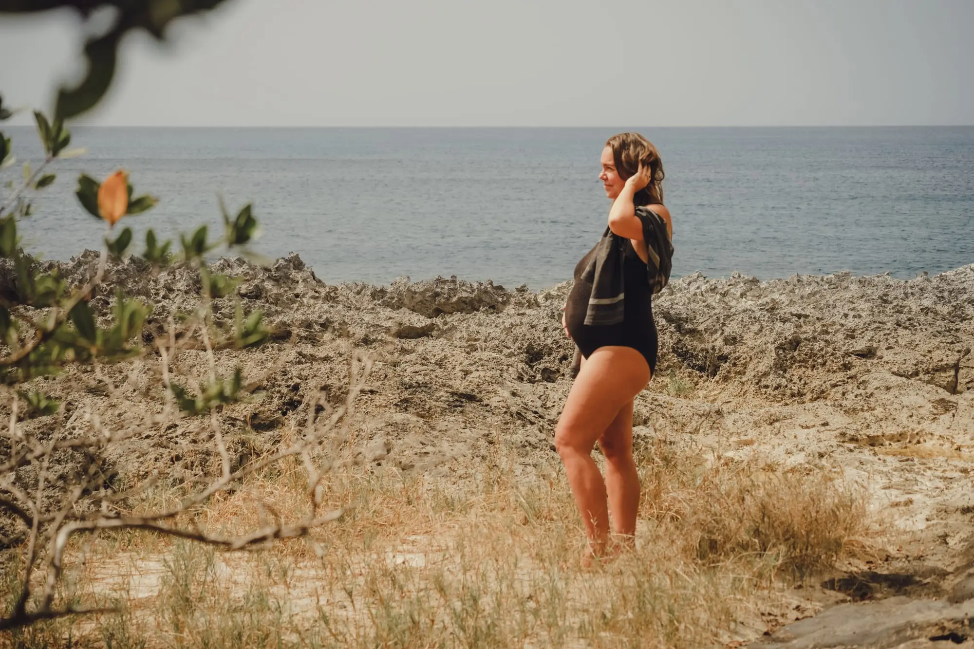 Séance grossesse à l'Anse Colas, à Anse-Bertrand : future maman de profil, une main dans les cheveux, sur les rochers qui bordent la mer.