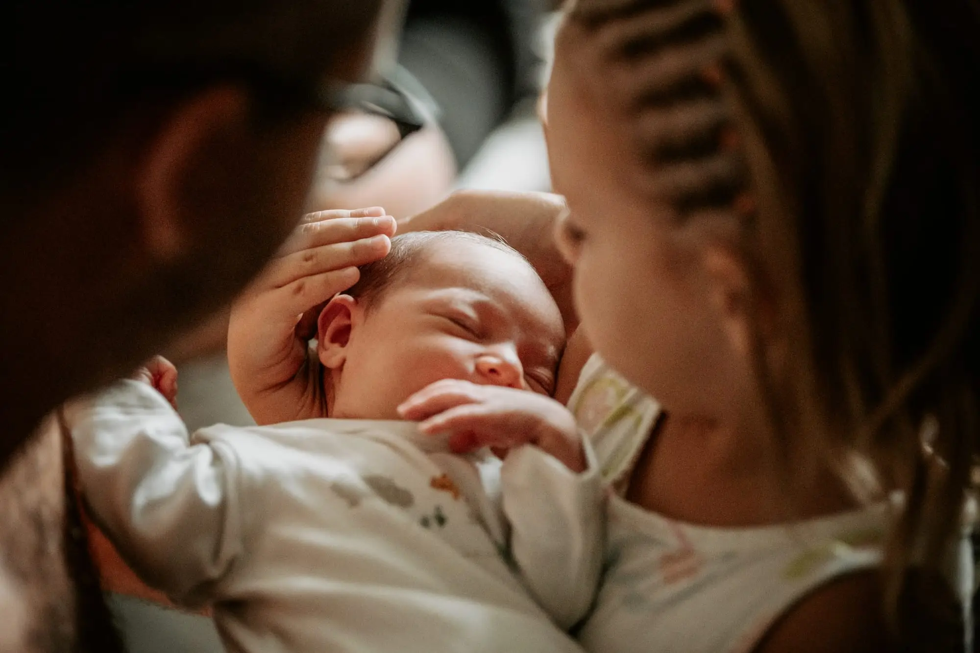Séance naissance à domicile, à Sainte-Anne : nouveau-né endormi dans les bras de ses parents, la main de son aînée posée sur son front.