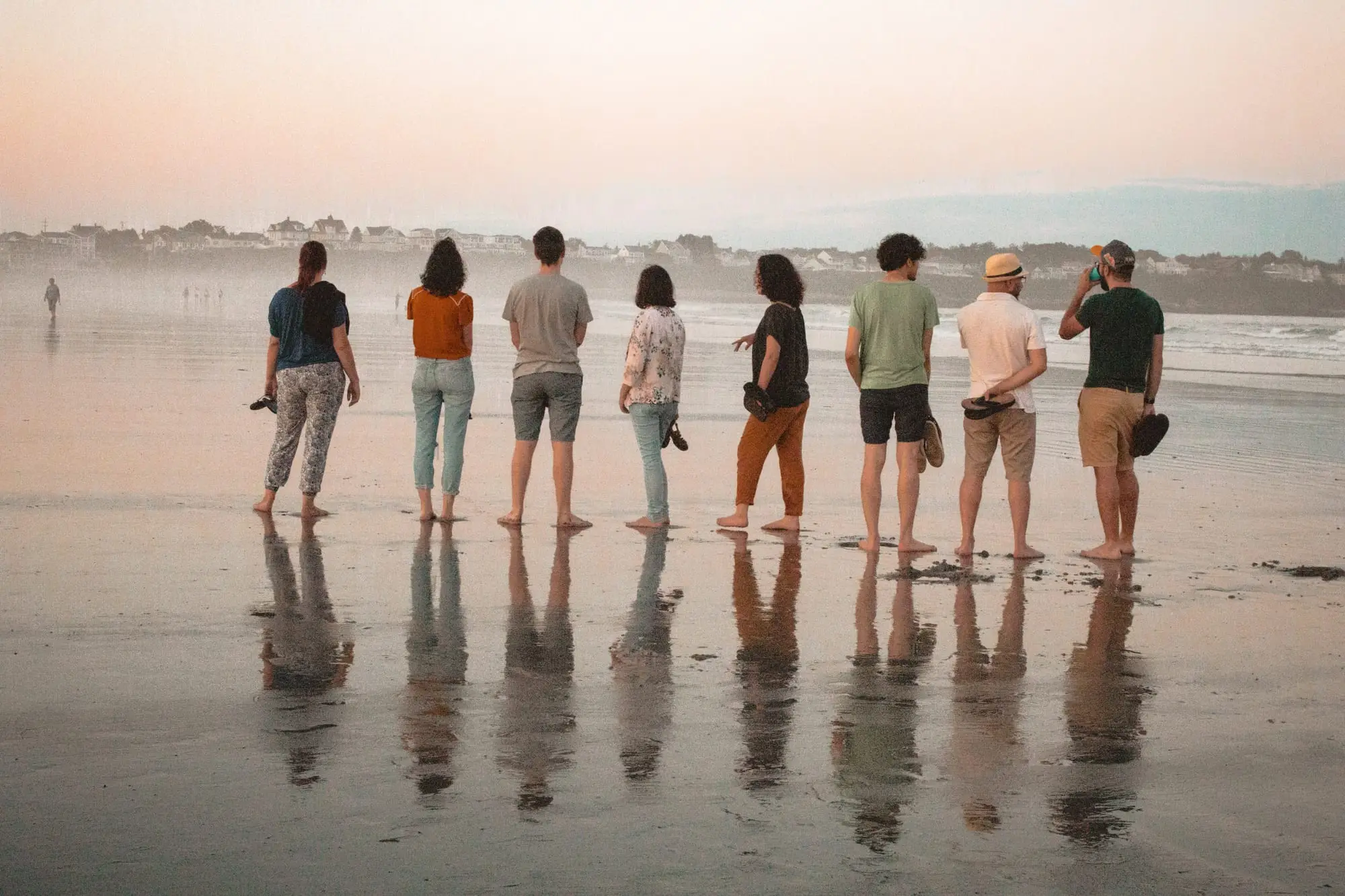 Séance entre amis sur la plage : toute la bande alignée de dos face à l'océan, pieds nus, les silhouettes reflétées dans le sable mouillé.