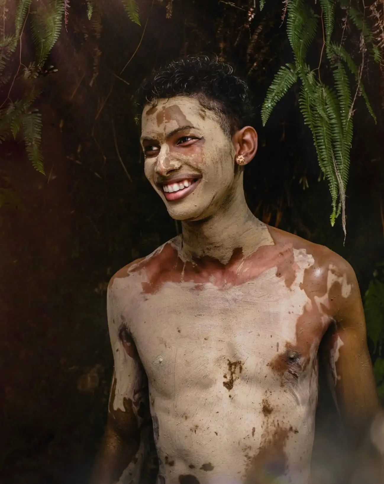 Séance portrait solo en rivière, à Petit-Bourg : un jeune homme couvert d'argile, souriant sous les fougères.